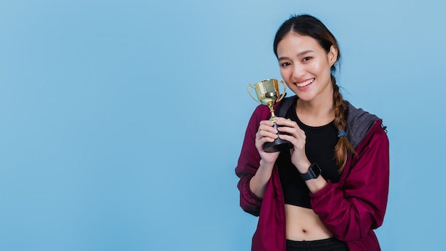 Portrait of confident beautiful asian fitness woman standing after winning and holding trophy on blue