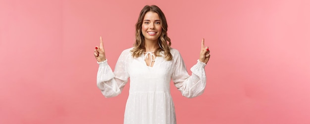 Portrait of confident beautiful young blond woman in white cute dress pointing fingers up at top advertisement looking at camera with beaming smile standing pink background