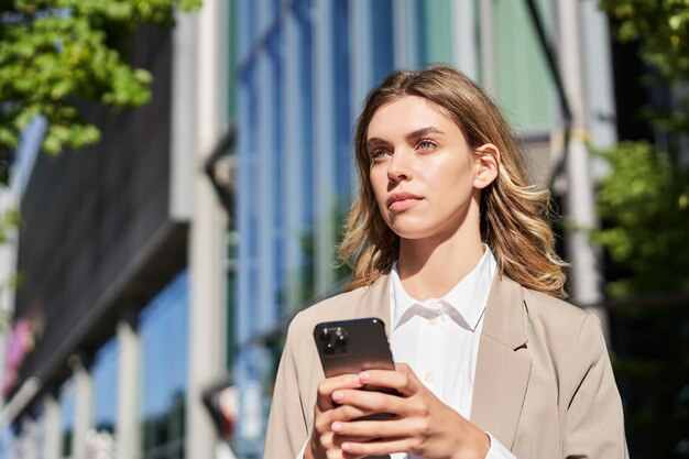 Portrait of confident businesswoman standing on street near office building using mobile phone order