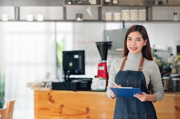 Portrait of confident female barista standing in front of the counter. Woman cafe owner in apron looking at camera and smiling.