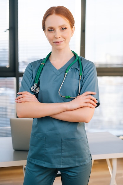 Портрет уверенно женщина-врач в сине-зеленой униформе, стоя на фоне окна в солнечный день в офисе медицинской клиники.