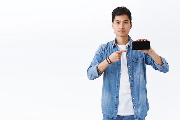 Portrait of confident good-looking asian male showing app or promo of company on smartphone screen, pointing at mobile phone recommending download