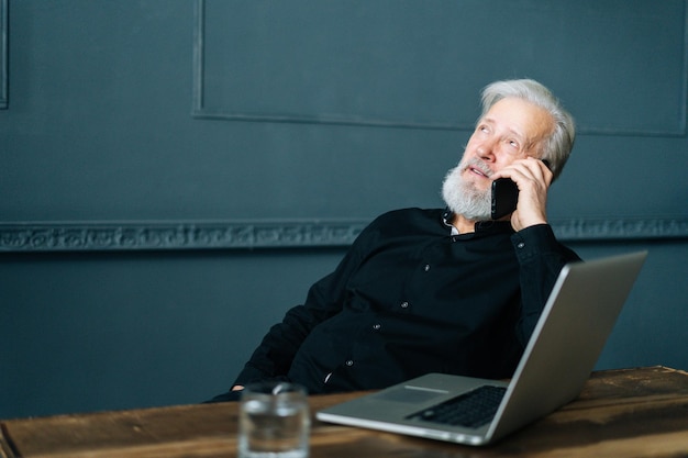 ラップトップコンピューターで木製のテーブルに座って携帯電話で話している自信を持って白髪のシニア大人のビジネスマンの肖像画。スマートフォンで会話をしているリラックスした成熟した年上の男性。