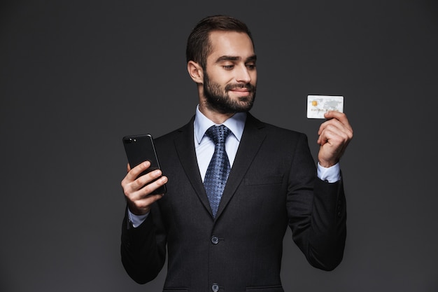 Portrait of a confident handsome businessman wearing suit isolated, using mobile phone, showing plastic credit card