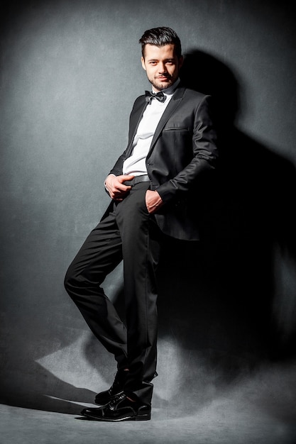 Portrait of confident handsome man in black suit with bowtie posing in dark studio background