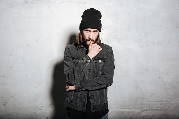 Portrait of a confident hipster guy in hat standing and looking at front isolated on wall