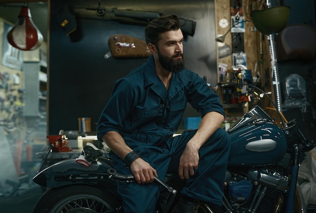 Portrait of confident man mechanic wearing overalls holding wrench in hand while sitting on repaired motorcycle. Workshop service concept