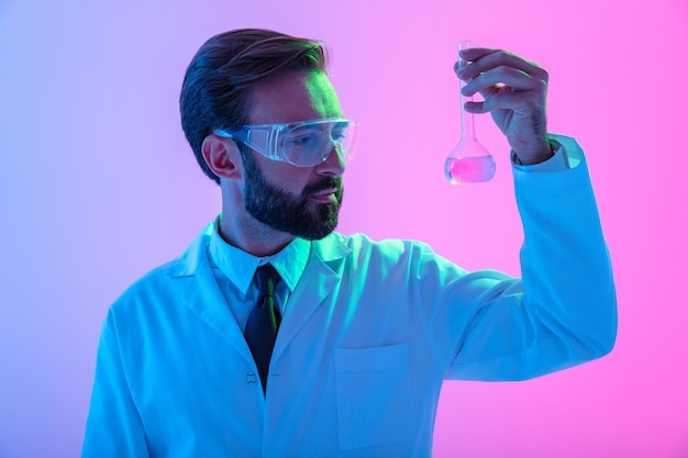 Photo portrait of a confident man scientist wearing unifrom and glasses standing isolated over blue-pink haze б looking at a flask with liquid