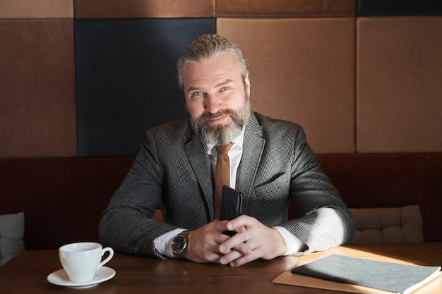 Portrait of confident mature businessman looking at camera sitting at the table with cup of coffee in cafe