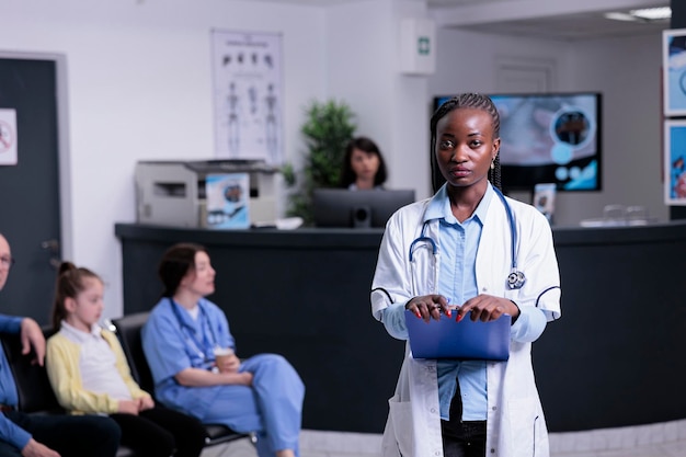 임상 상담을 위해 환자 예약을 기다리는 병원 리셉션에 서 있는 자신감 있는 의료진의 초상화. 의료 데이터 클립보드를 들고 병원 유니폼에 아프리카계 미국인 의사.
