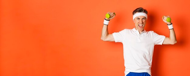 Portrait of confident middleaged fitness guy doing sports over orange background flex biceps and hol