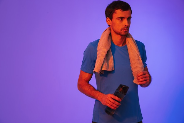 Ritratto di uno sportivo motivato fiducioso in piedi isolato sul muro viola, che riposa dopo l'allenamento, bevendo acqua