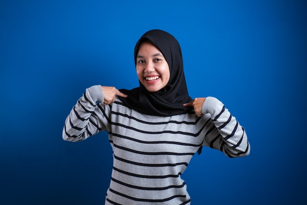 Portrait of confident muslim lady wearing hijab smiling and pointing at her self, against blue background