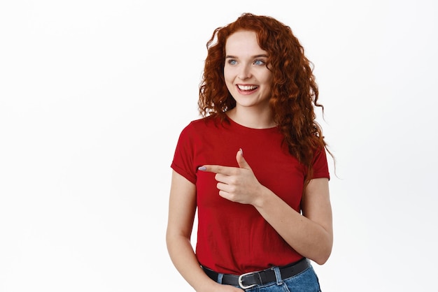 Portrait of confident redhead female student with curly long hair pointing and looking left at banner logo showing advertisement white background
