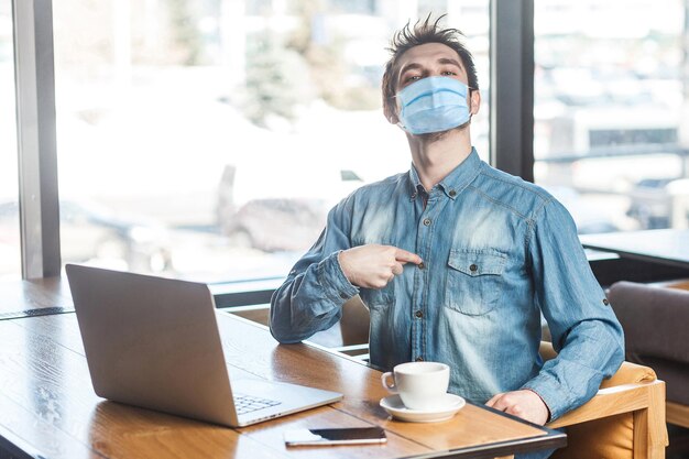 Portrait of confident satisfied egoistic young man with surgical medical mask in blue shirt sitting, working on laptop and pointing finger at himself with proud. indoor working and health care concept