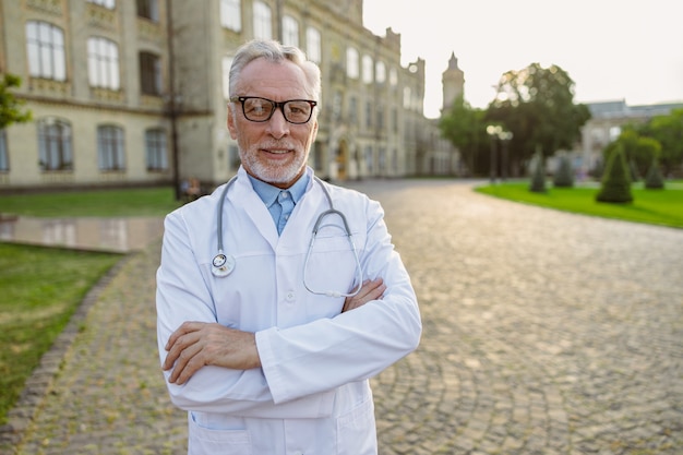 Portrait of confident senior male doctor in lab coat and glasses smiling at camera standing with