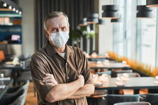 Portrait of confident senior restaurant manager in mask and brown shirt standing with crossed arms against rows of tables