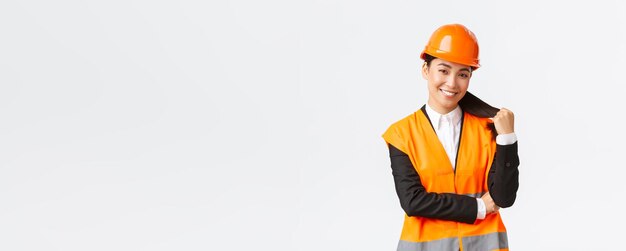 Portrait of confident smiling asian female construction manager engineer at building area wearing safety helmet and reflective jacket looking delighted at project being pleased with finished work