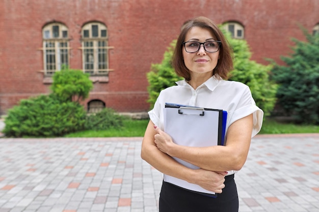 Portrait of confident smiling businesswoman female teacher counselor with clipboard paper documents Positive smiling woman copy space school building office building background