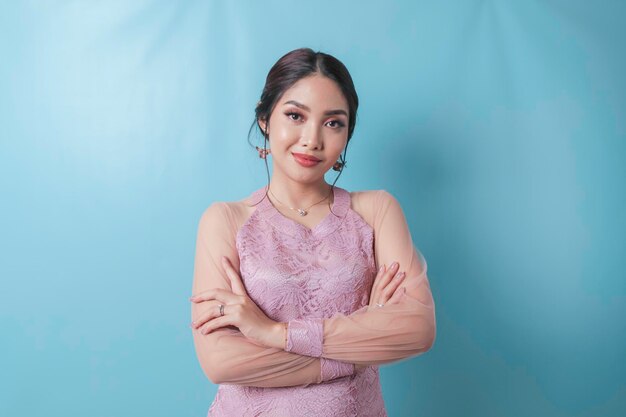 Portrait of a confident smiling girl standing with arms folded and looking at camera isolated over blue background wearing modern kebaya dress