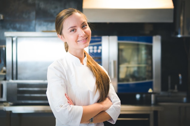 Il ritratto dello chef giovane donna sicuro e sorridente vestito in cucina professionale uniforme bianca è sullo sfondo