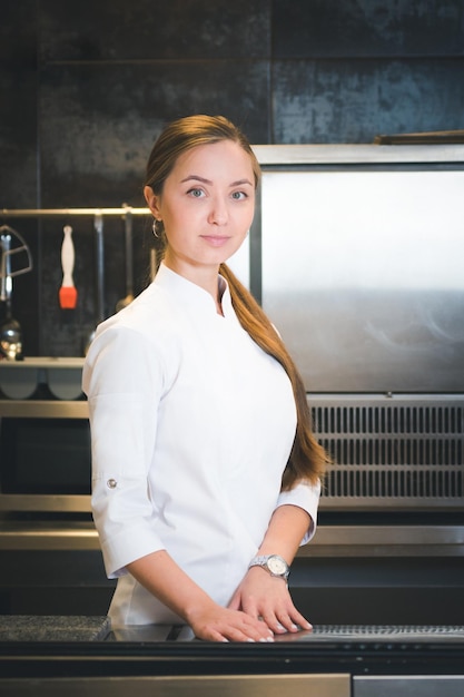 Портрет уверенной и улыбающейся молодой женщины-повара, одетой в белую униформу, профессиональная кухня на заднем плане