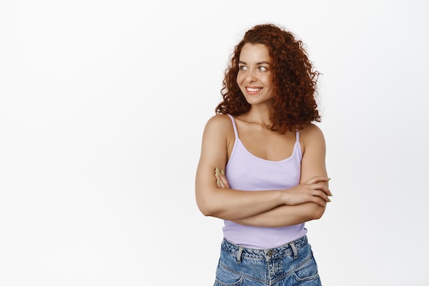 Portrait of confident, stylish redhead woman with curly hair, cross arms on chest and smiling happy, looking left at copy space, standing in tank top against white background