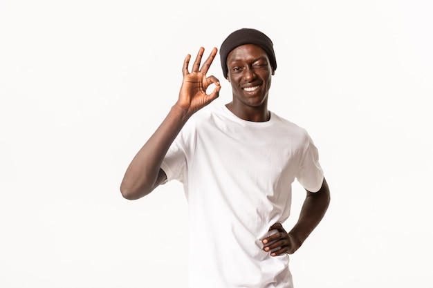 Portrait of confident and upbeat african-american cool guy in beanie, assure all good, winking in approval and showing okay gesture, guarantee quality