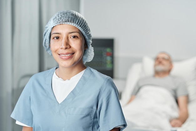 Portrait of confident young adult woman working in hospital smiling at camera her patient lying in bed on background