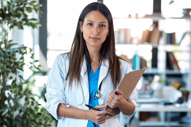 Photo portrait of confident young female doctor looking at camera while holding a clipboard and standing in the consultation.