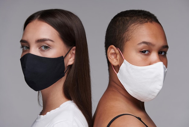 Portrait of confident young interracial women wearing cloth masks standing back to back against gray background