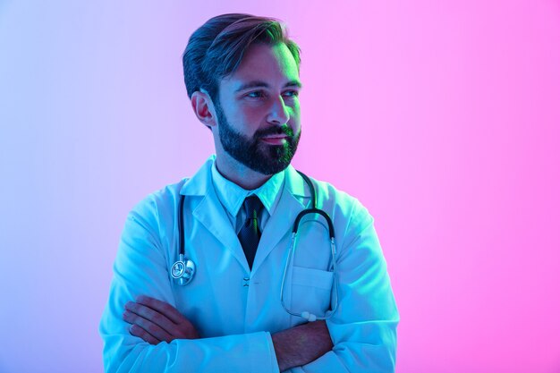 Portrait of a confident young man doctor wearing uniform standing isolated over pink-blue haze , arms folded, looking away