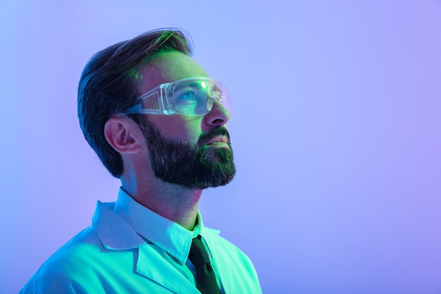 portrait of a confident young man scientist wearing uniform and glasses standing isolated over pink-blue haze , looking away