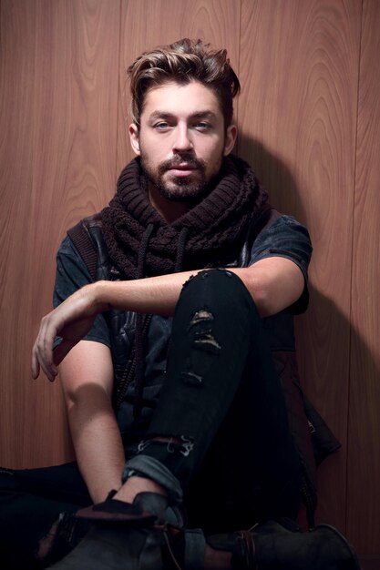 Photo portrait of a confident young man with a hair face sitting on a wooden wall looking at the camera