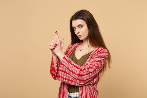 Portrait of confident young woman in casual clothes looking camera holding hands like gun isolated on pastel beige background in studio. People sincere emotions, lifestyle concept. Mock up copy space.