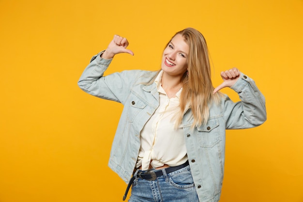 Ritratto di giovane donna sicura di sé in abiti casual in denim che guarda la fotocamera, puntando i pollici su se stessa isolata su uno sfondo giallo arancione della parete in studio. concetto di stile di vita delle persone. mock up spazio di copia.