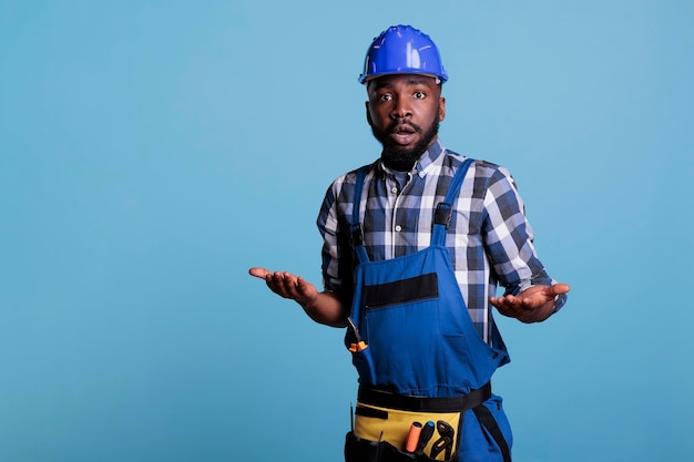 파란색 배경에 작업복을 입고 포즈를 취한 혼란스러운 아프리카계 미국인 건축업자의 초상화가 불확실한 표정을 짓고 있습니다. 어깨를 으쓱하는 남자, 스튜디오 샷 나는 개념을 모른다.