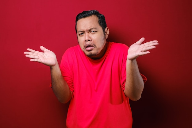 Portrait of confused annoyed man shrug raising arms, asking and having no idea what happening over red background