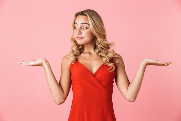 Portrait of a confused beautiful cute woman in dress posing isolated over pink wall showing copyspace