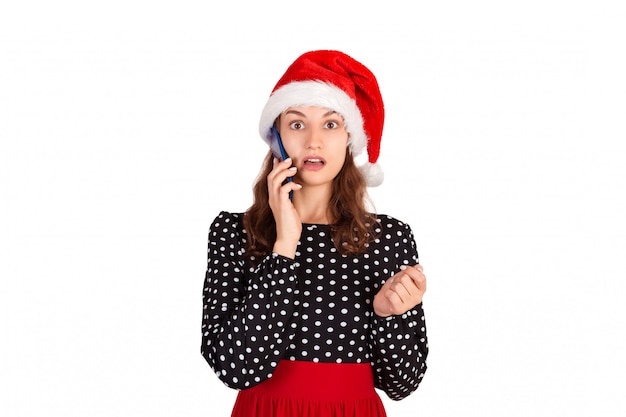 Portrait of a confused cute girl in dress talk at mobile phone. emotional girl in santa claus christmas hat isolated 