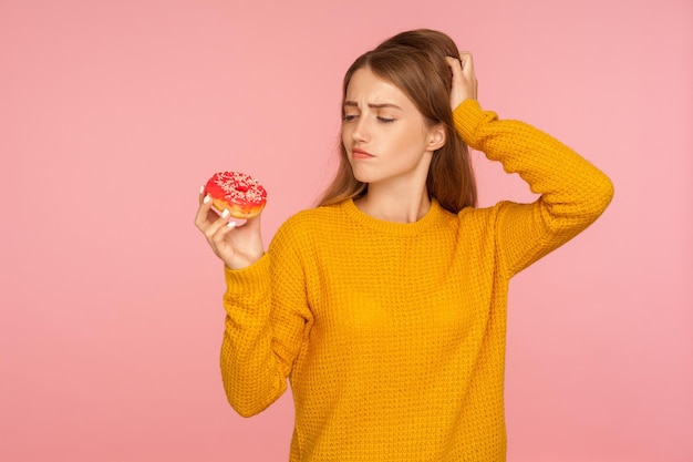 Portrait of confused ginger girl in sweater scratching head and hesitating to eat sweet doughnut wants sugary donut for snack and doubting about junk food thinking over unhealthy food studio shot