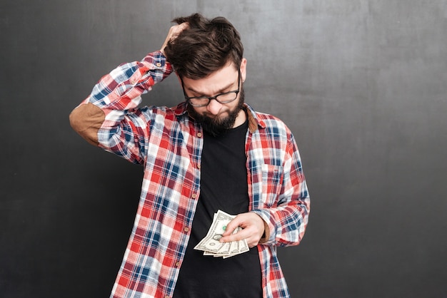 Foto ritratto di un uomo confuso vestito con una camicia in una gabbia e con gli occhiali in piedi sulla lavagna mentre tiene soldi