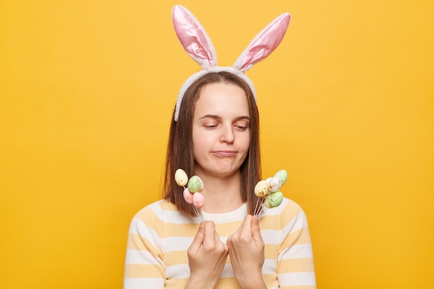 Foto ritratto di donna confusa e perplessa che indossa orecchie da coniglio che tiene la torta di pasqua pop isolata su sfondo giallo che decide di mangiare o dessert non zuccherato