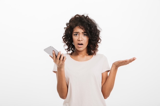 Portrait of confused puzzled woman with afro hairstyle wearing t-shirt holding cell phone and expressing misunderstanding, isolated over white wall