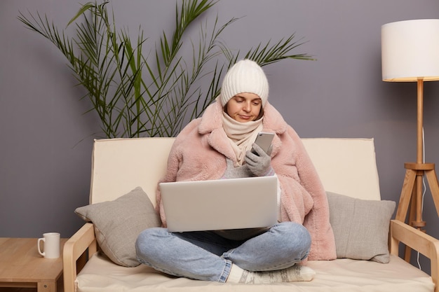 Portrait of confused sad woman wearing cap and winter coat sitting on the sofa at home in cold room working on portable computer and using mobile phone trying to call heating service