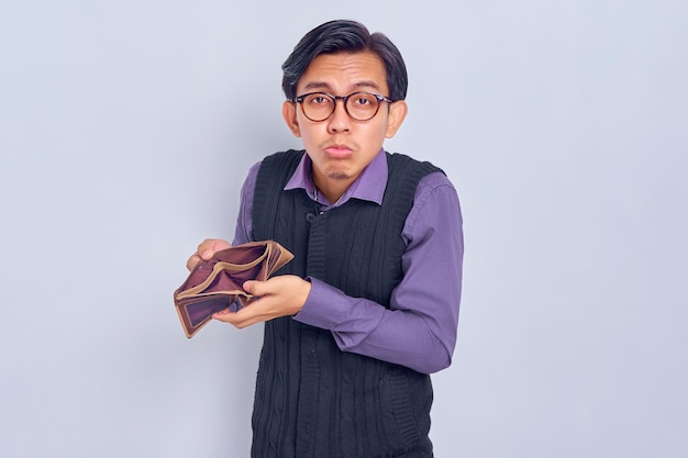 Portrait of confused and sad young Asian man in 30s wearing shirt and vest showing at camera brown empty wallet isolated on white background