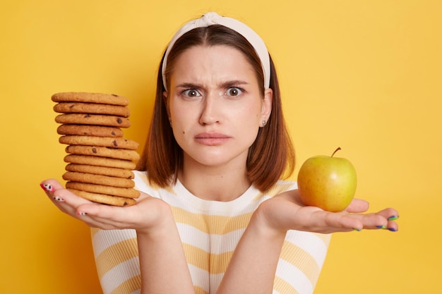 Portrait of confused woman wearing striped shirt and hair band looking at camera with puzzlement holding cookies and apple does not know what to choose posing isolated over yellow background