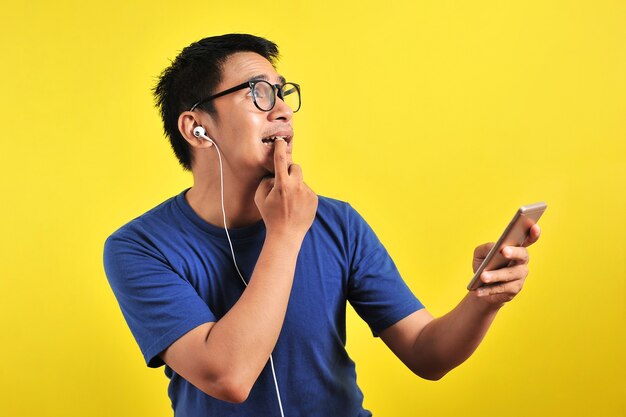 Portrait of confused young asian man bitting his finger and listening live music from smartphone with headset, isolated on yellow background