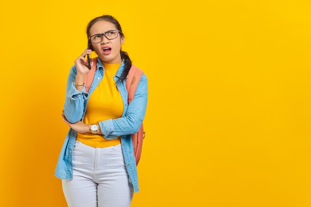 Portrait of confused young Asian woman student in casual clothes with backpack talking on smartphone and looking at copy space isolated on yellow background. Education in college university concept