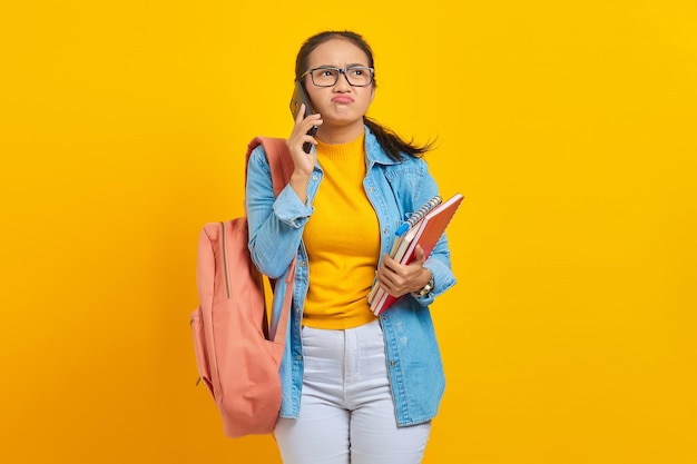 Portrait of confused young Asian woman student in denim clothes with backpack talking on mobile phone and holding book isolated on yellow background